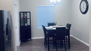 Un comedor con una mesa y sillas, una araña, un reloj en la pared y una ventana con persianas.