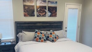 Un dormitorio con una cama, dos mesitas de noche, una ventana con persianas y tres cuadros colgados en la pared.