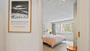 Een slaapkamer met een groot raam, een lijst met een afbeelding aan de muur, een bed met een roze deken, en een houten stoel.