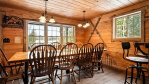L’intérieur d’un chalet en bois, avec une table à manger entourée de chaises, de grandes fenêtres et un lustre.