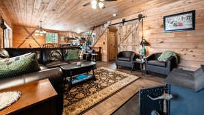 Un intérieur de chalet chaleureux avec un plafond en bois, un ventilateur de plafond, une cheminée, un coin repas et un salon comprenant un canapé et une table basse.
