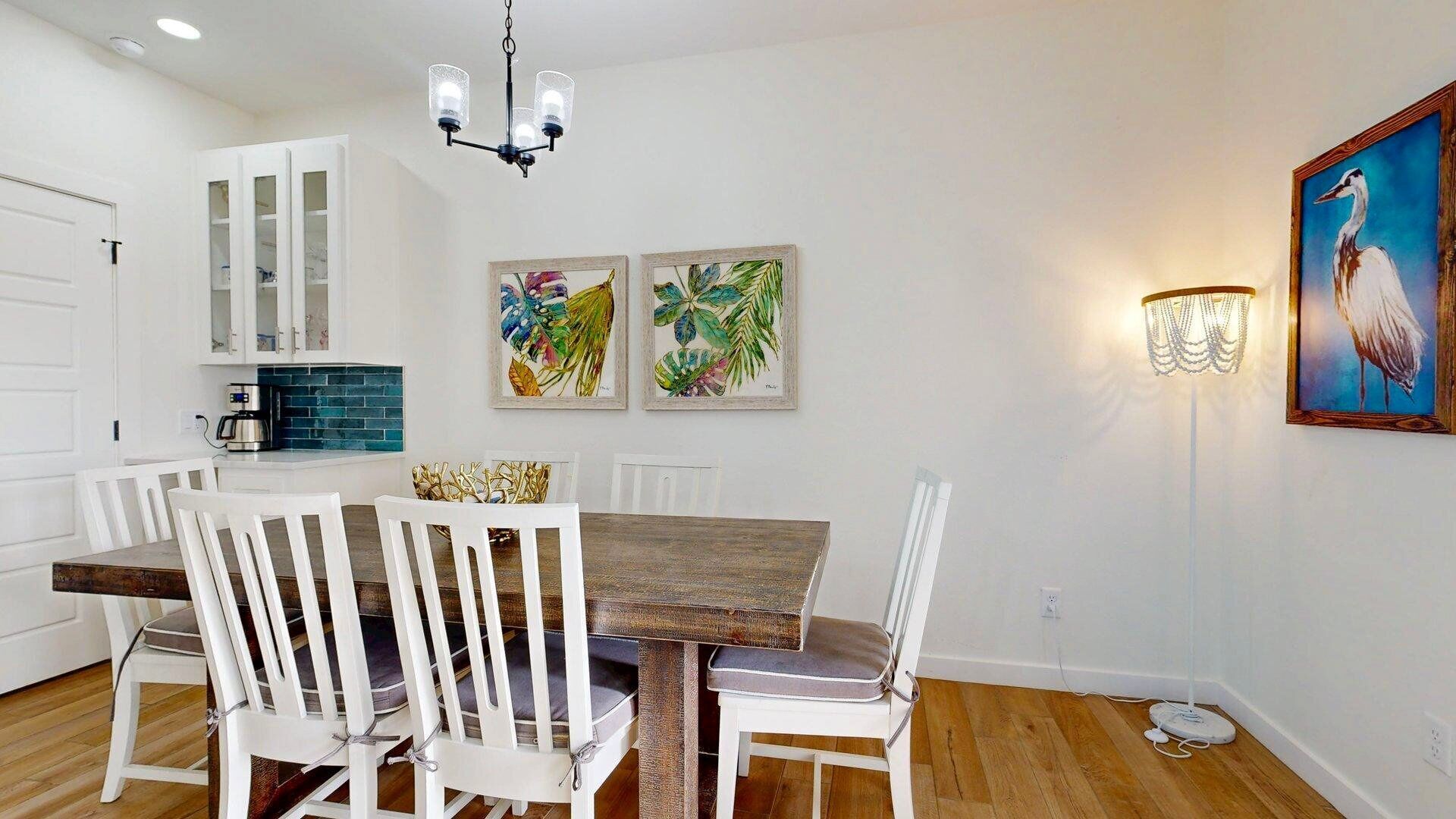 Un comedor con una mesa de madera y sillas blancas, una cocina con armarios blancos y un salpicadero azul, y arte mural que representa aves y follaje tropical.