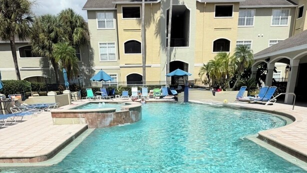 Piscina cristalina com espreguiçadeiras e guarda-sóis, cercada por um edifício com varandas.