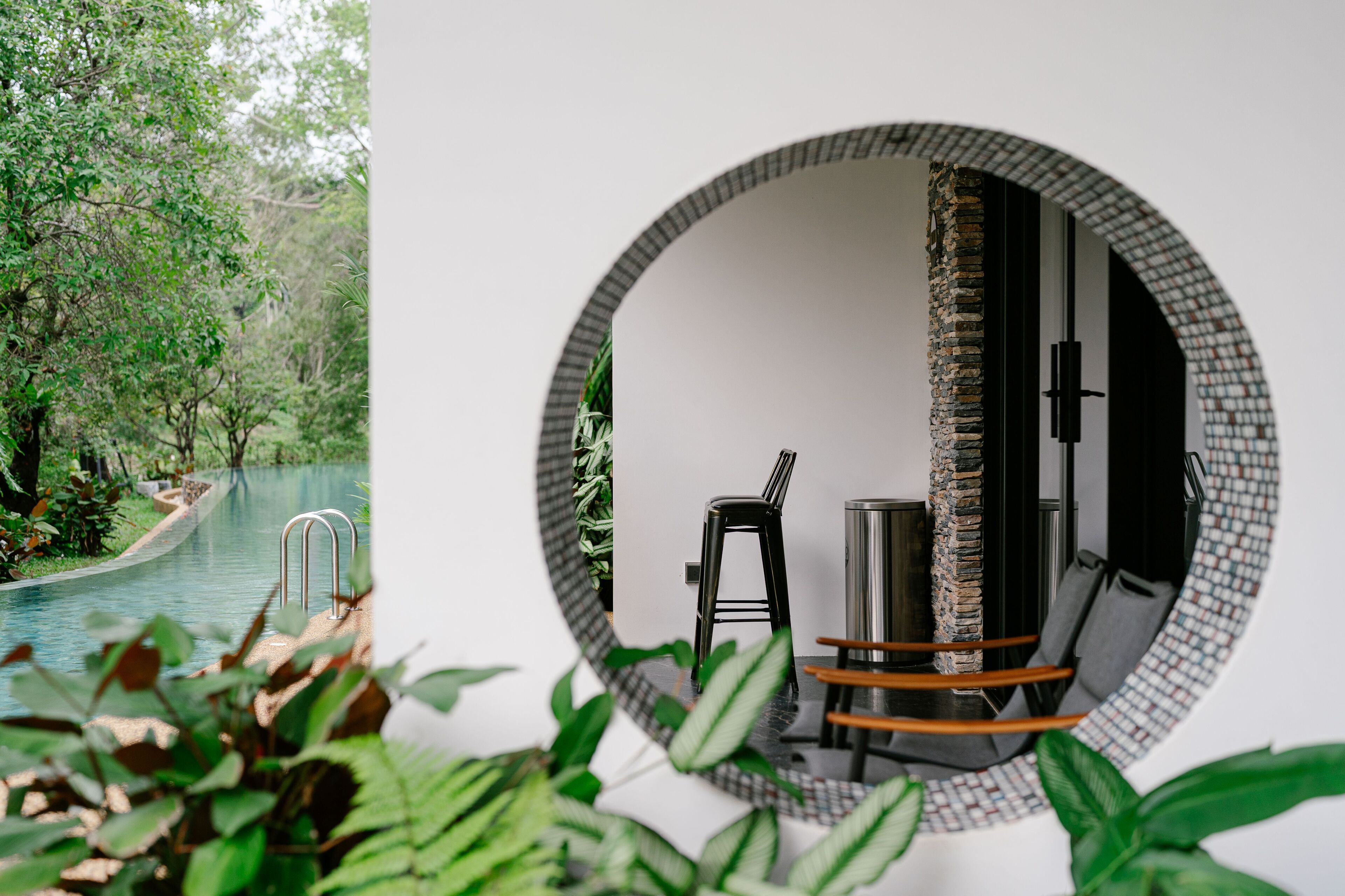 A circular mirror reflecting a pool and a chair, surrounded by greenery.
