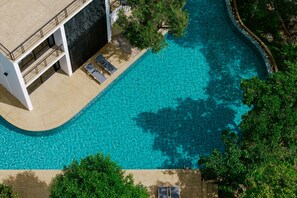 An aerial view of a modern hotel with a large swimming pool, surrounded by lush greenery and a multi-story building with balconies.