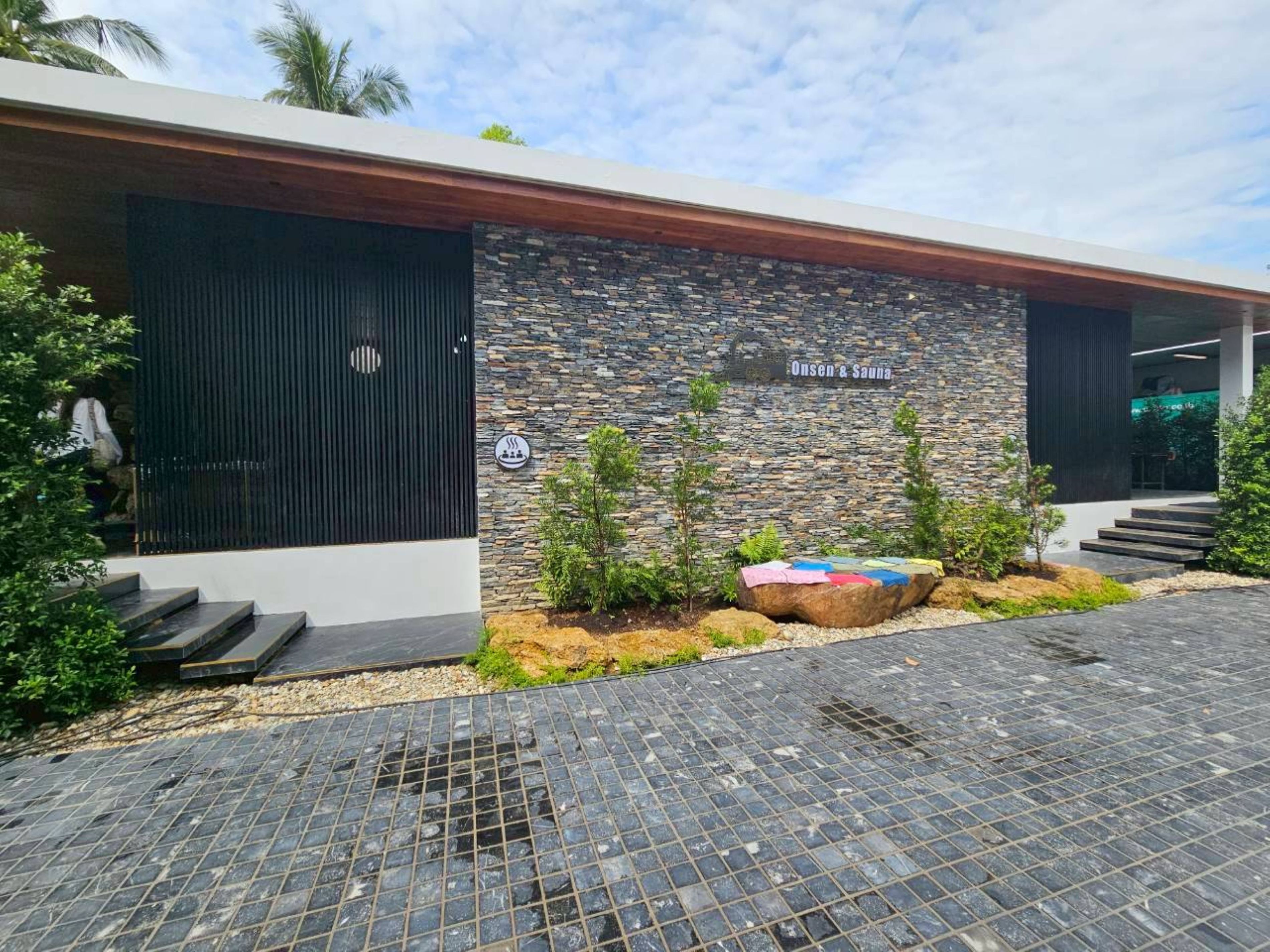 A modern building with a stone facade, a sign reading "Onsen & Sauna," and a small garden area with plants and a colorful rock.
