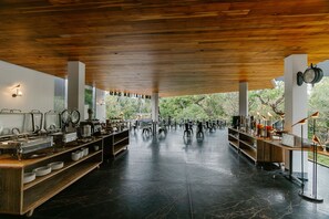 A spacious indoor area with a polished black floor, wooden ceiling, and white walls. There are multiple tables and chairs arranged for dining, with a buffet setup on the left side.