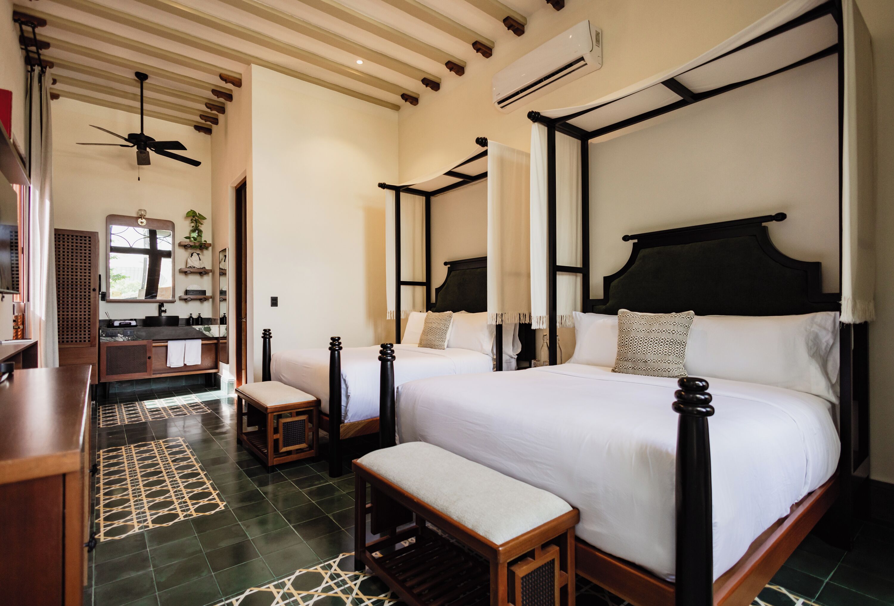 Une chambre d’hôtel avec un lit à baldaquin, un coin salon avec un banc, une coiffeuse avec un miroir et un ventilateur de plafond.