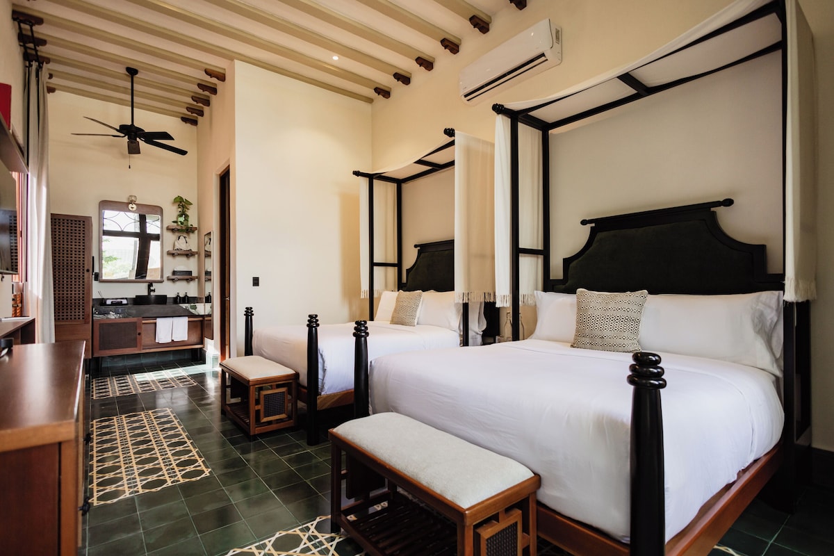 A hotel room with a four-poster bed, a sitting area with a bench, a vanity with a mirror, and a ceiling fan.