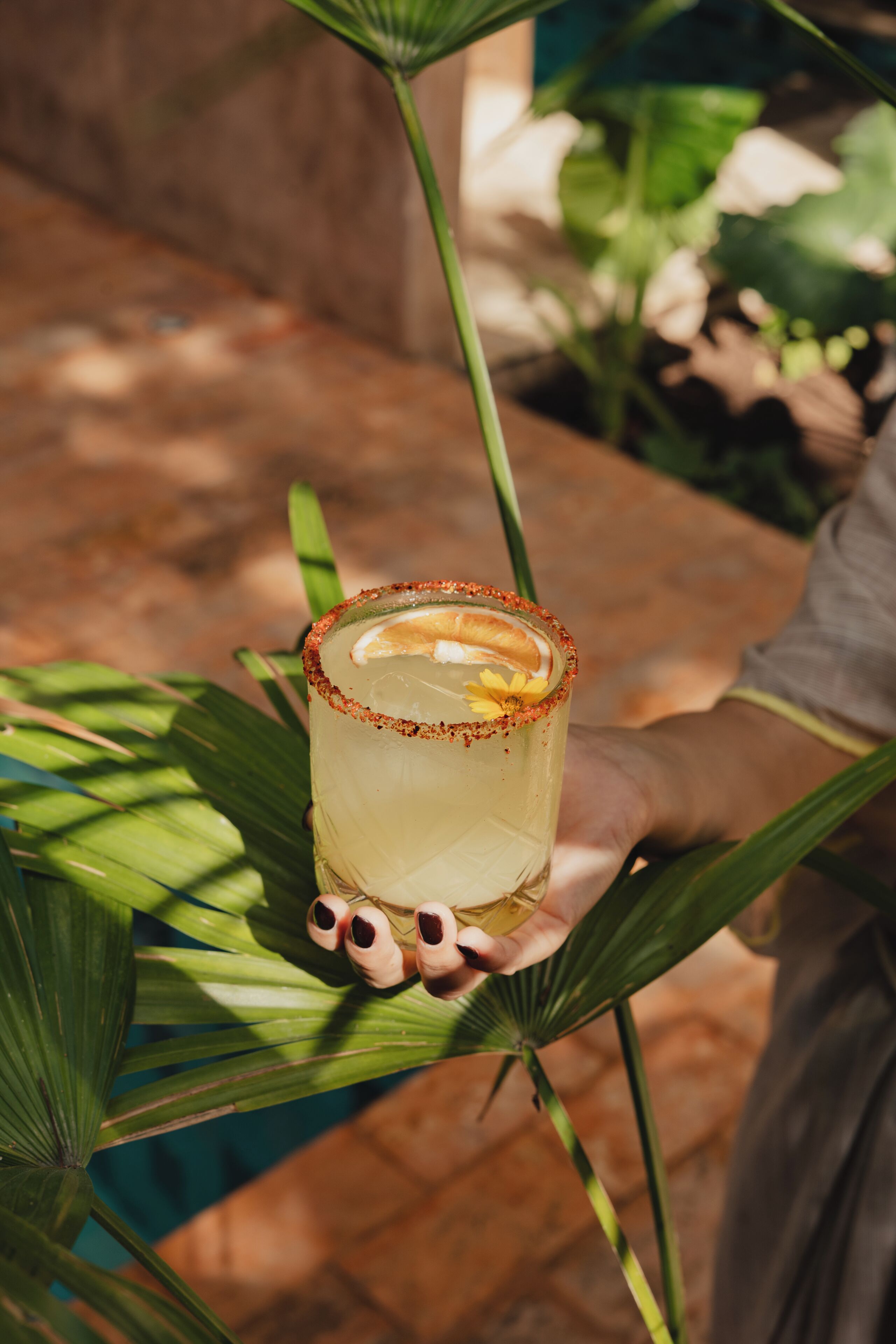 A hand holding a glass with a rim of red flakes, containing a yellowish liquid and a slice of orange.