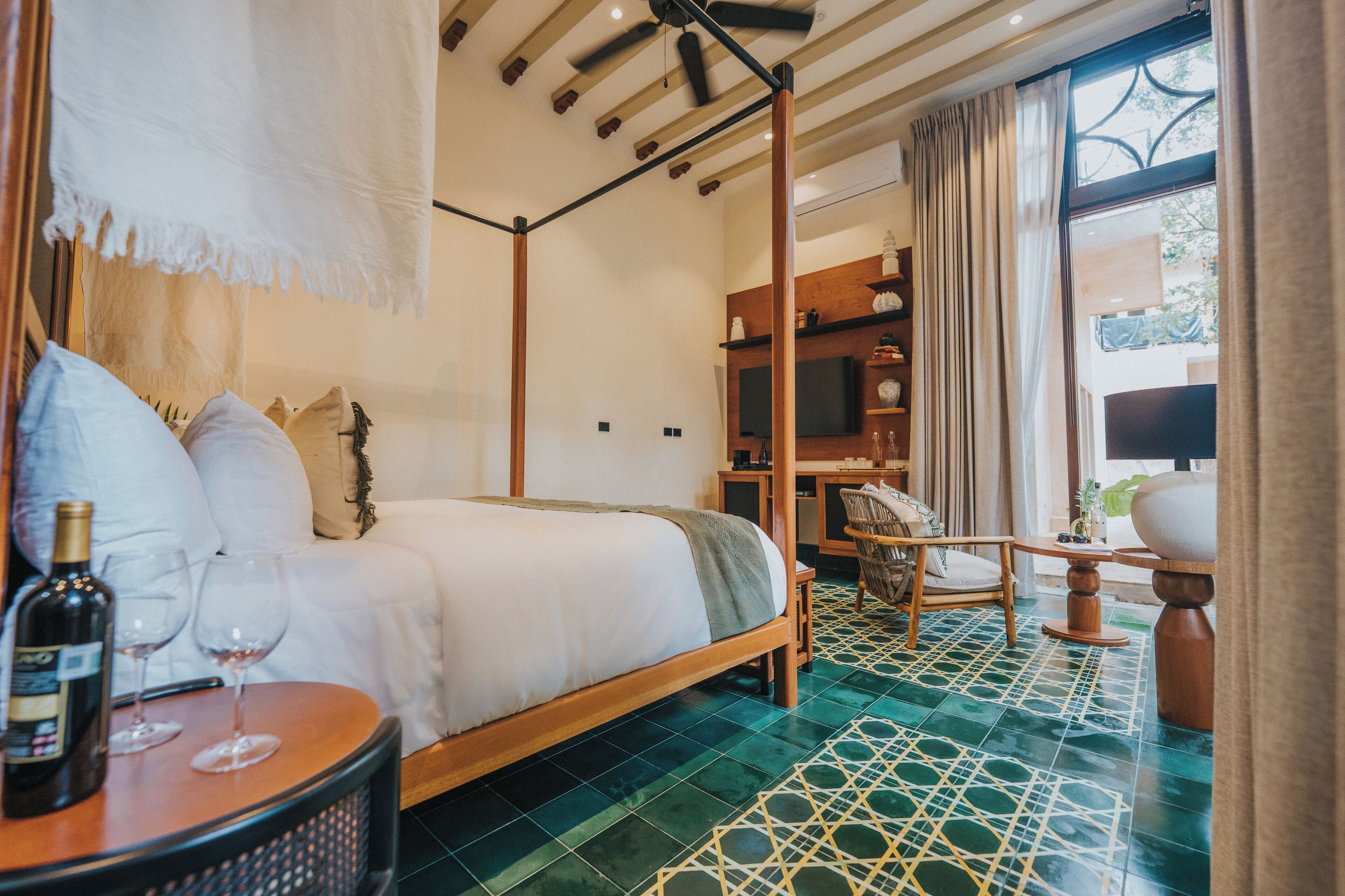 A four-poster bed with a wooden frame, a bedside table with a bottle and glasses, a chair, and a TV on a wooden shelf.