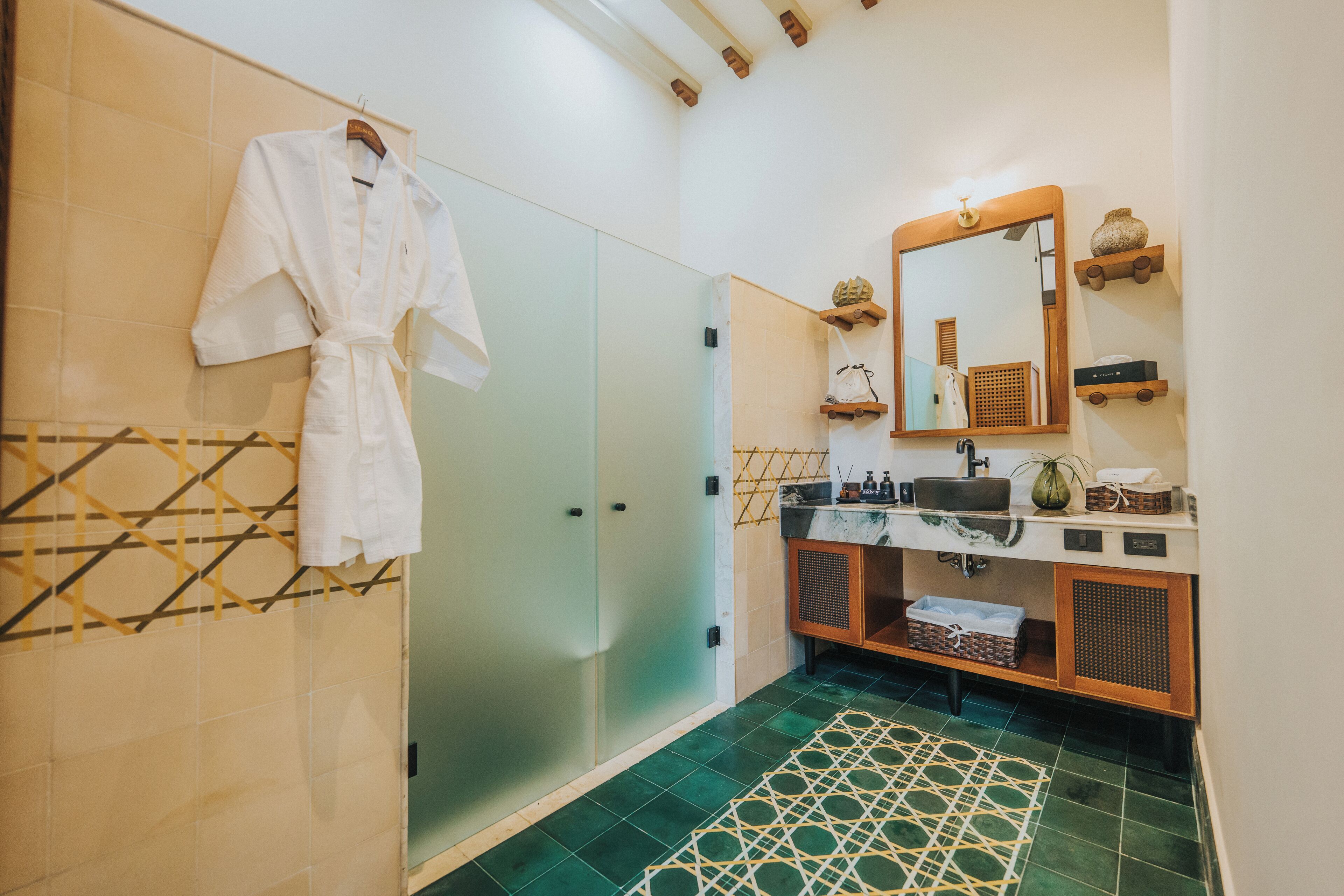 A modern bathroom with a glass shower enclosure, a white robe hanging on the wall, a wooden vanity with a mirror, and a patterned floor.