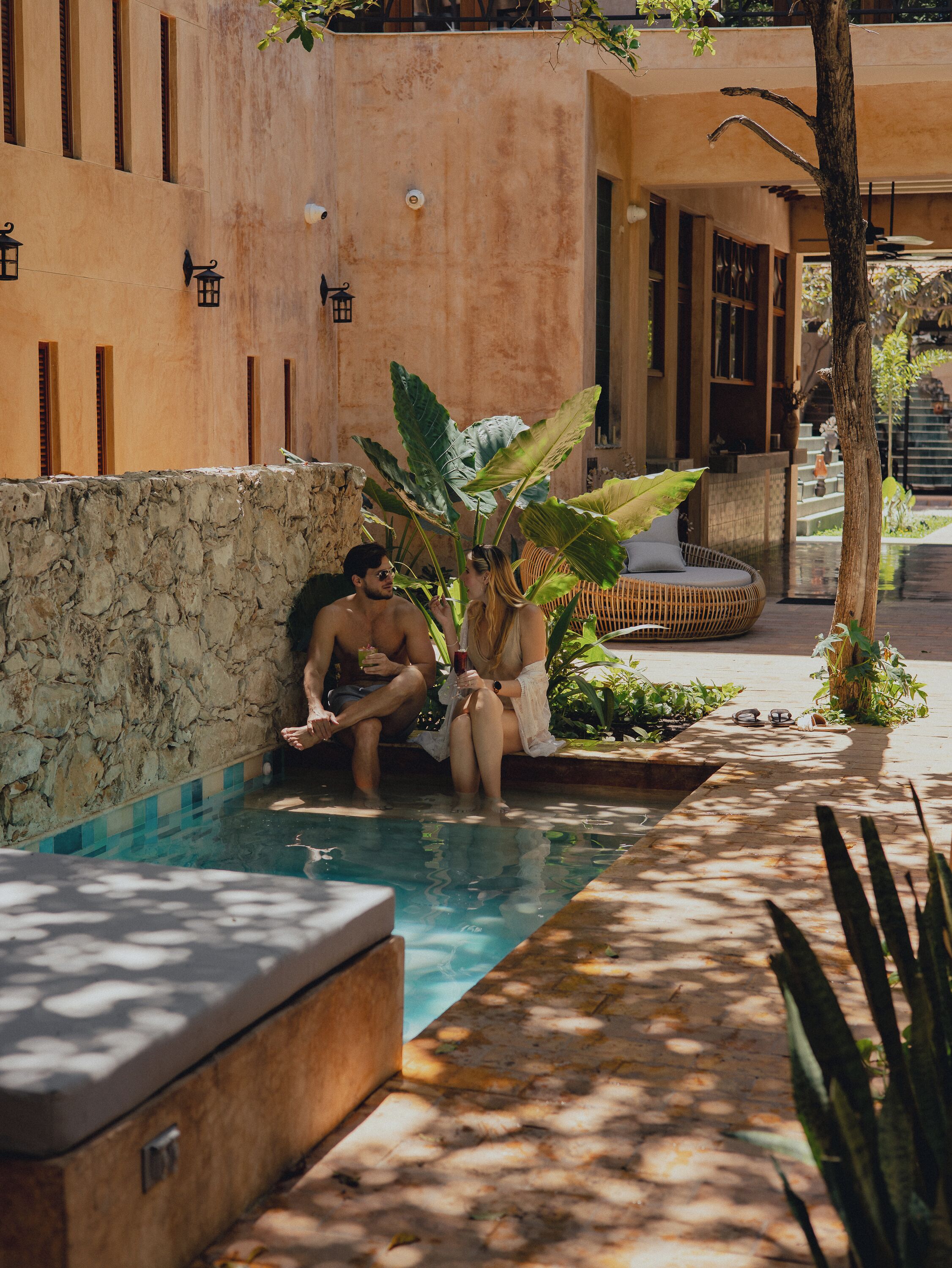 A pool area with a stone wall, a lounge chair, and a large plant.