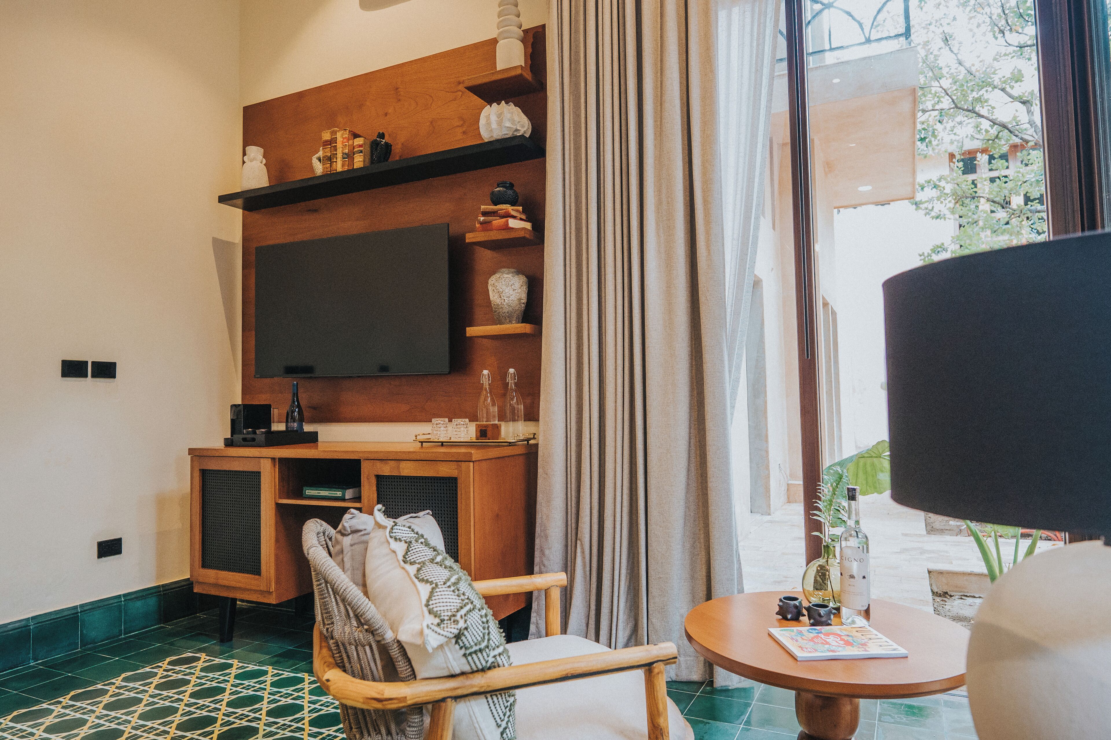 A cozy room with a wooden entertainment unit, a flat-screen TV, a comfortable chair, a small round table with a magazine, and a view of the outdoors through a window with curtains.