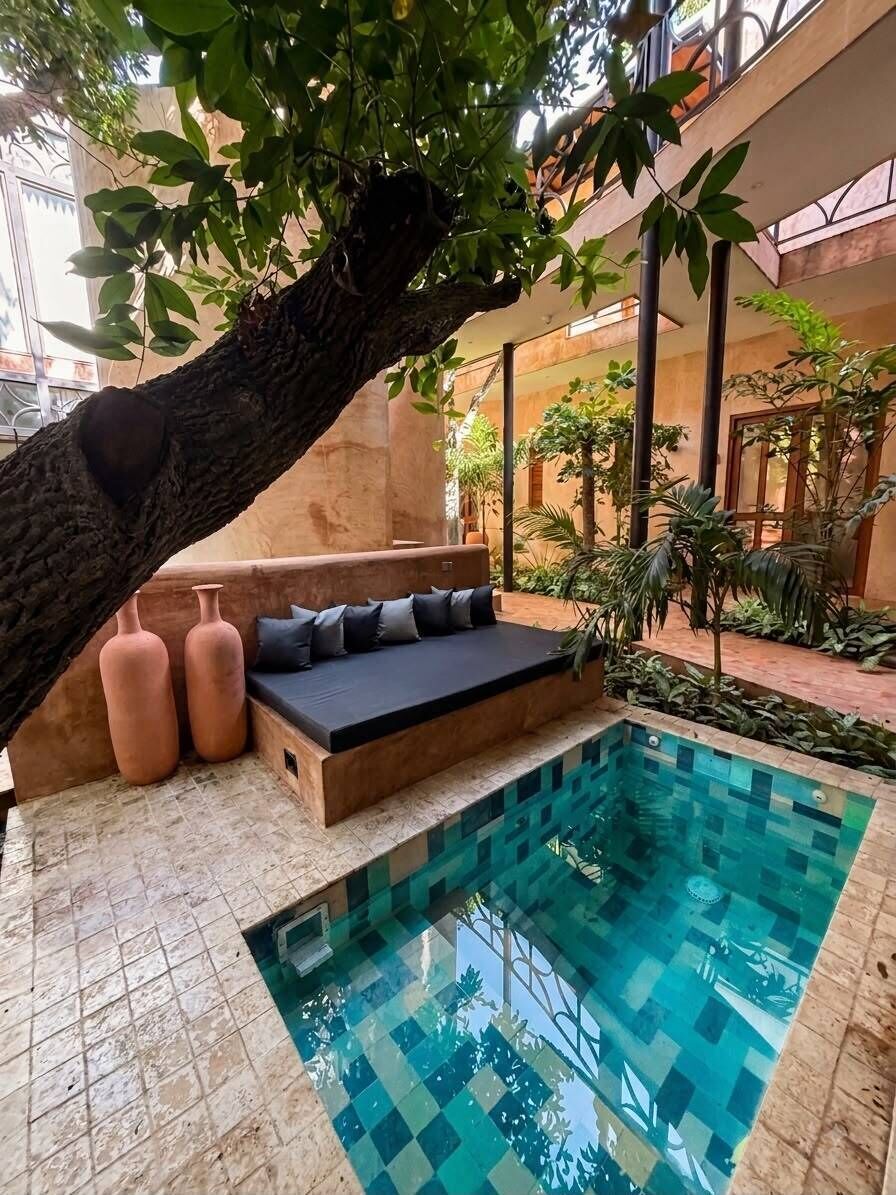 Un espace piscine avec un coin salon aménagé sous un arbre, entouré de plantes et doté d’un sol carrelé.