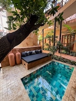 A pool area with a seating area under a tree, surrounded by plants and a tiled floor.