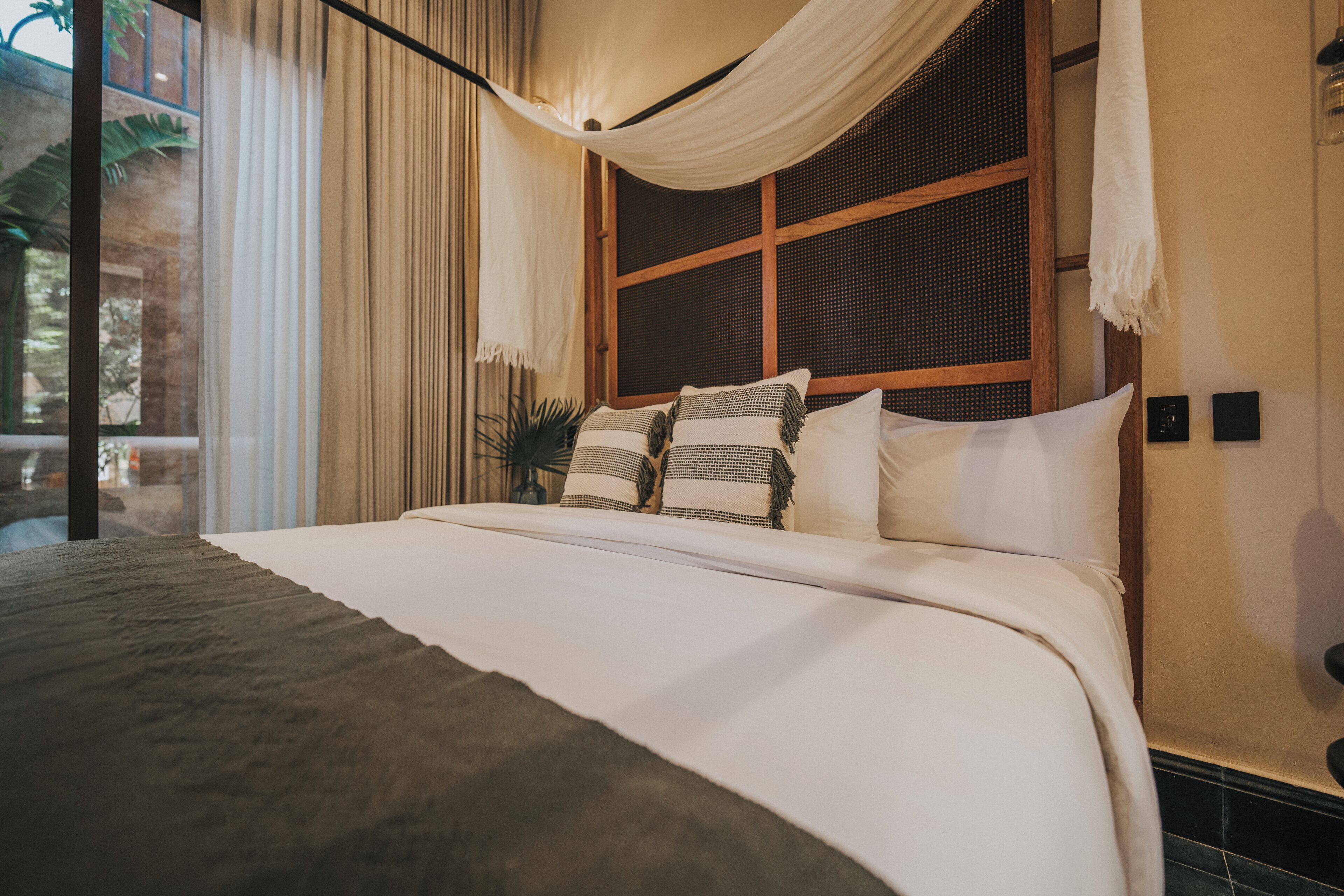 A neatly made bed with white linens and multiple pillows, a large window with curtains, and a view of greenery outside.
