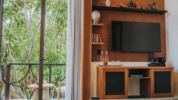 A room with a large flat-screen TV mounted on a wooden wall, a wooden console table with books and decorative items, and a view of lush greenery through a glass door.