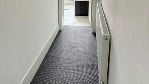 Un pasillo estrecho con un radiador a la derecha que conduce a un baño con bañera y ventana.