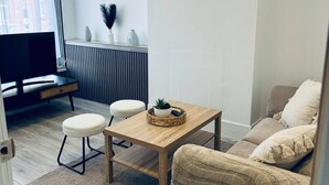 Una sala de estar moderna con una mesa de madera, dos otomanas blancas, un sofá beige y un televisor sobre un mueble negro.