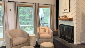 A cozy living room with a fireplace, two armchairs, ottomans, and a view of the outdoors. - Harbor Springs Adventure Base—Beach, Trails & Skiing (Harbor Springs)