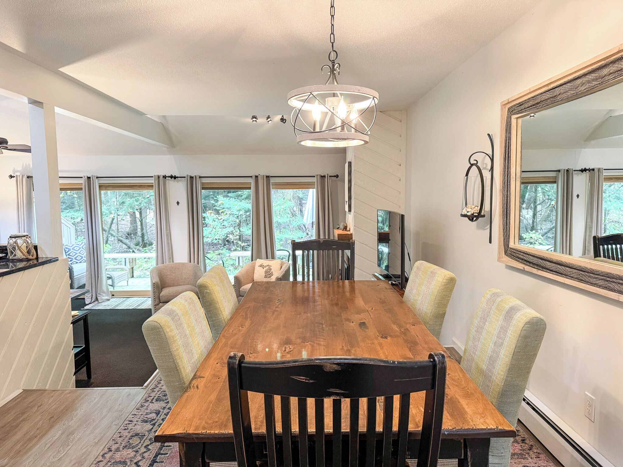 A dining area with a wooden table, chairs, and a chandelier. There is a large mirror on the wall and a view of the outdoors through large windows.