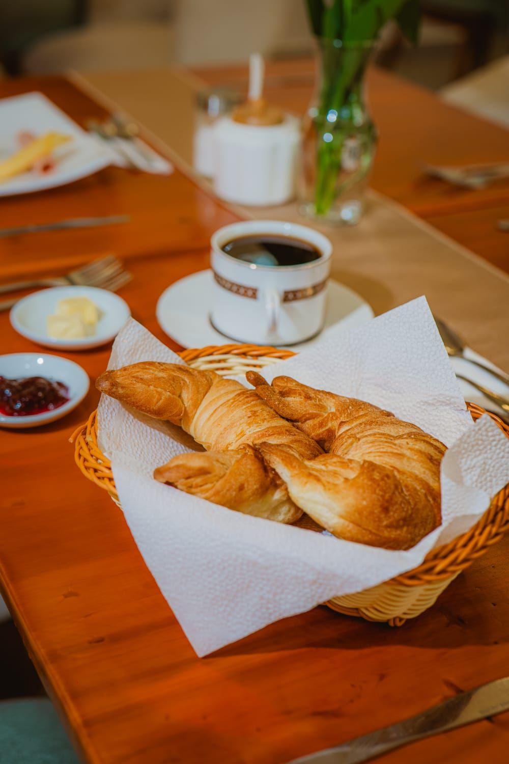 A table with a basket of croissants, a cup of coffee, and small dishes of butter and jam.