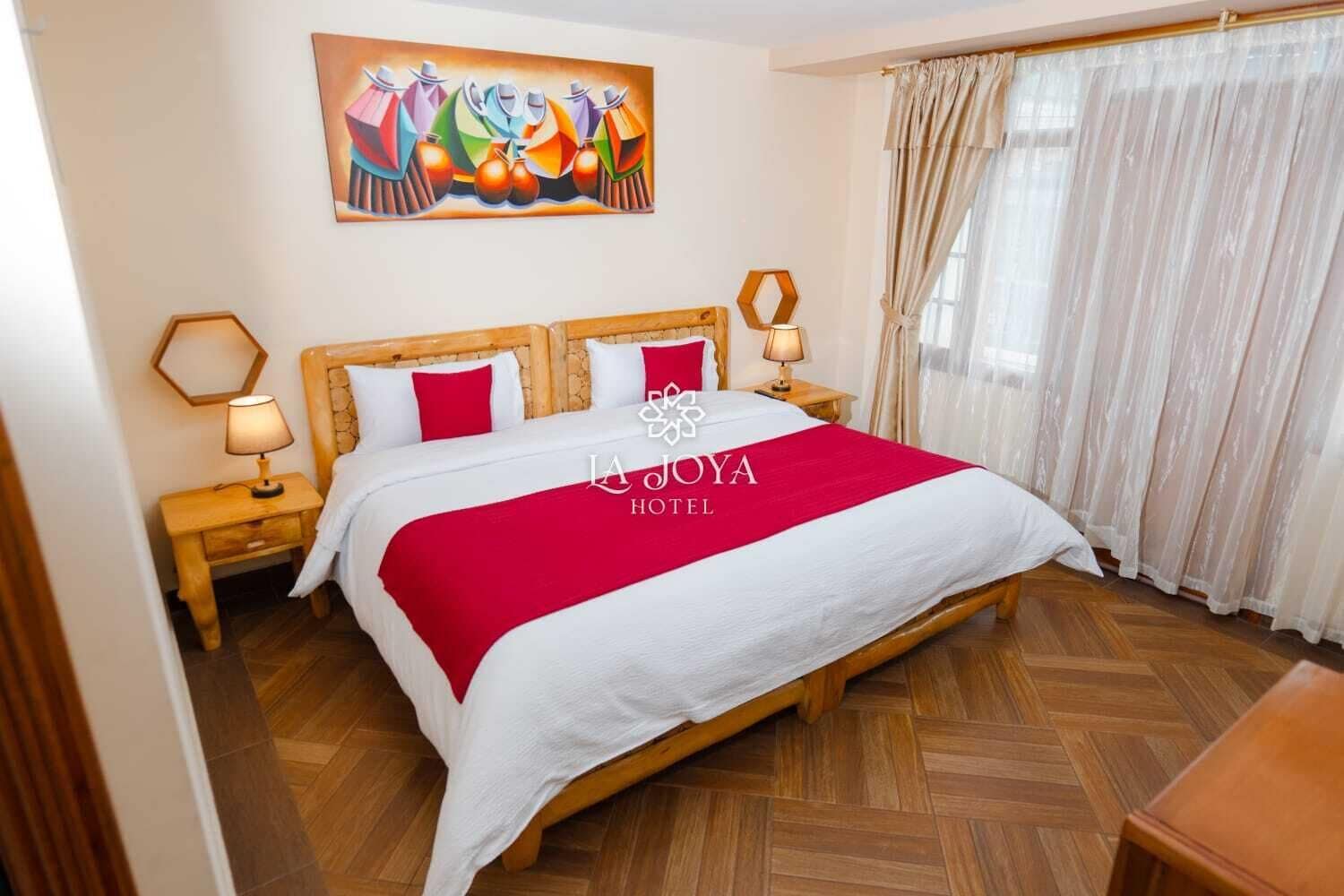 Habitación de hotel con una cama grande, cabecera de madera, dos mesitas de noche con lámparas, una colcha de color rojo y blanco, un cuadro abstracto colorido sobre la cama y una ventana con cortinas traslúcidas.