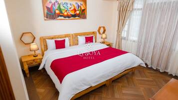 Habitación de hotel con una cama grande, cabecera de madera, dos mesitas de noche con lámparas, una colcha de color rojo y blanco, un cuadro abstracto colorido sobre la cama y una ventana con cortinas traslúcidas.