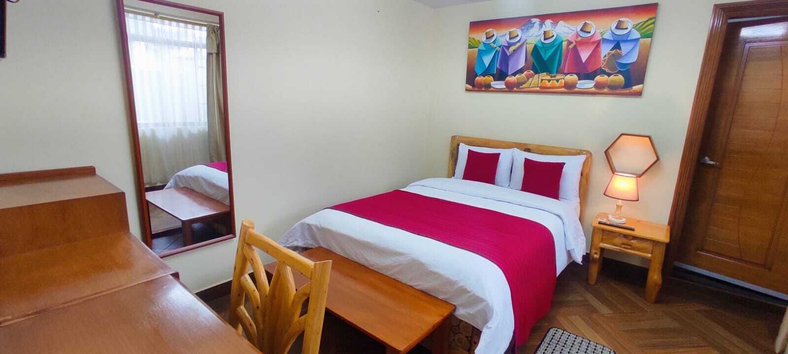 A bedroom with a bed, a desk, a chair, a nightstand with a lamp, and a painting on the wall.