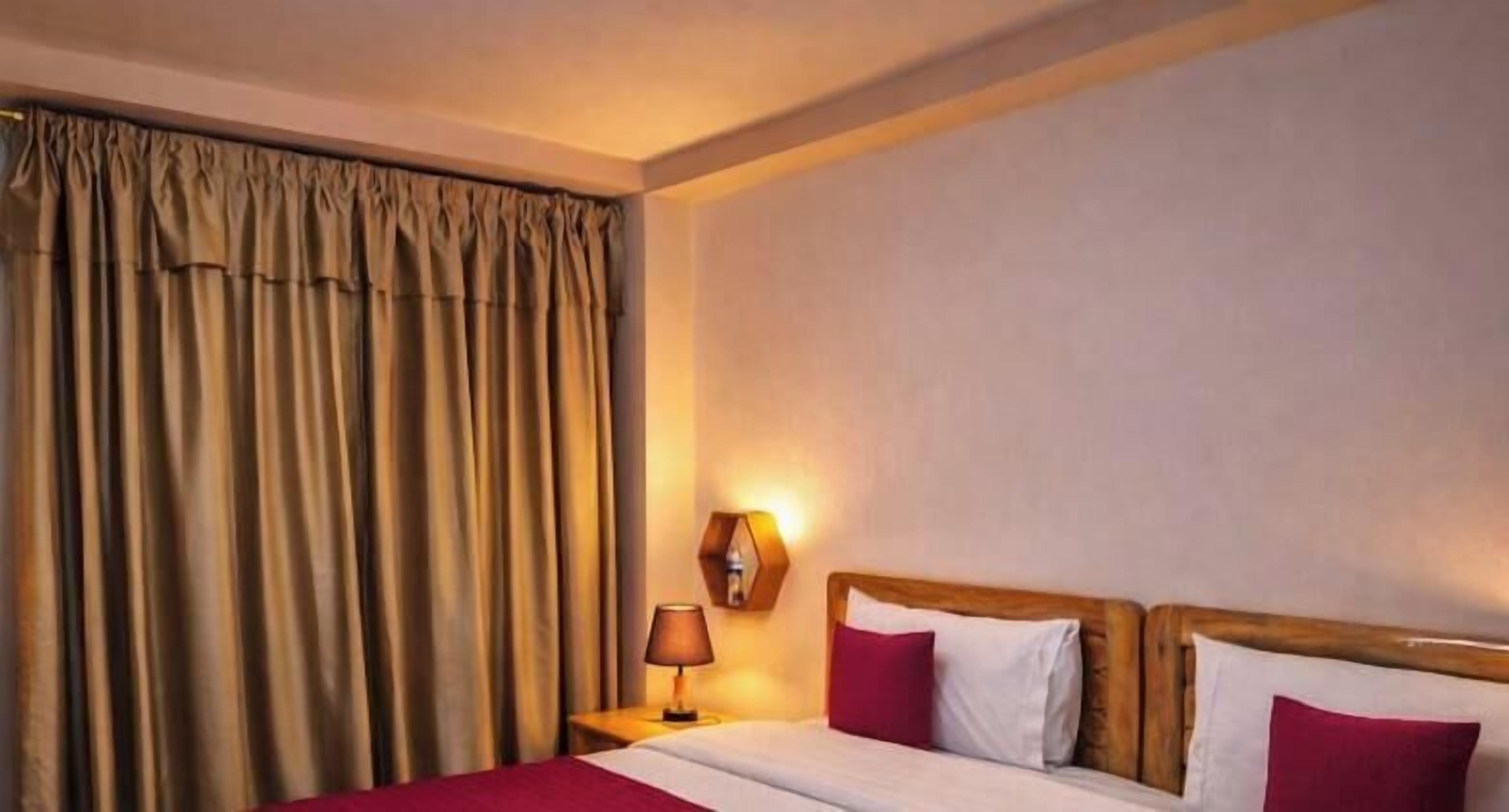 A hotel room with a bed, curtains, a nightstand with a lamp, and a wall-mounted light fixture.