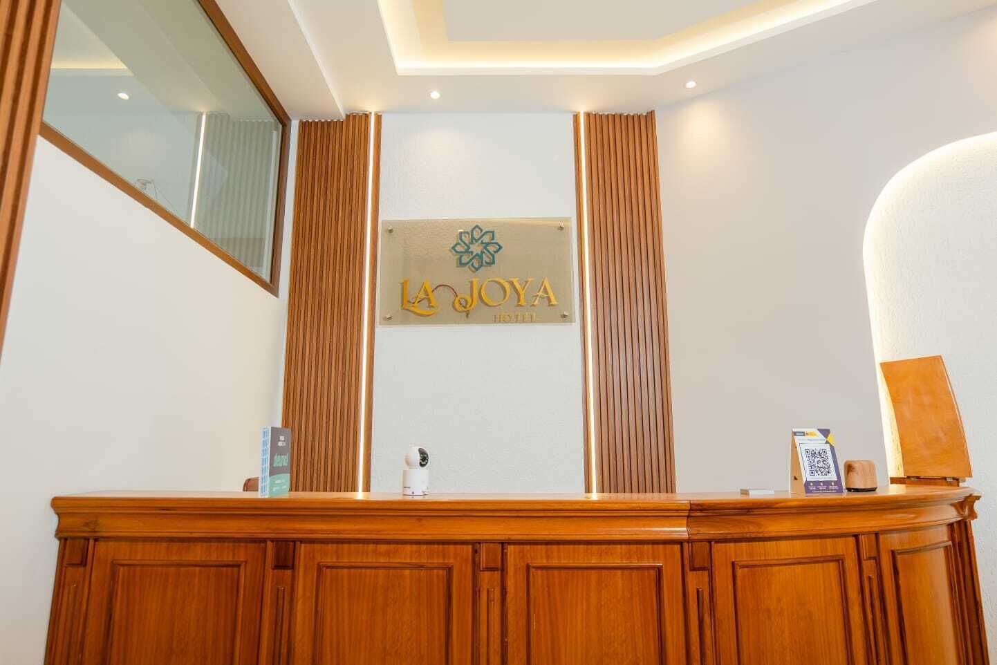 Un comptoir d’accueil avec un revêtement en bois, une enseigne au-dessus indiquant « HÔTEL LA JOYA », et une caméra de surveillance placée sur le comptoir.