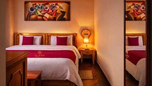 A hotel room with a bed, a desk, a nightstand with a lamp, and a painting on the wall. - HOTEL LA JOYA (Otavalo)