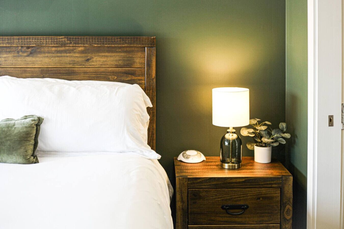 A neatly made bed with white linens and a green pillow, a bedside lamp, a small white vase with a plant, and a wooden nightstand with a drawer.