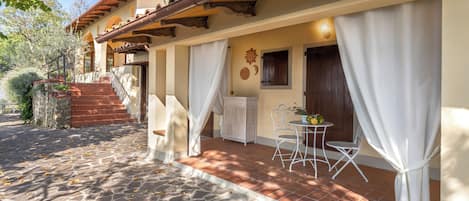 Une terrasse dallée de pierres, avec une fenêtre ornée d’un rideau blanc, une petite table avec des chaises et un escalier menant à l’entrée.
