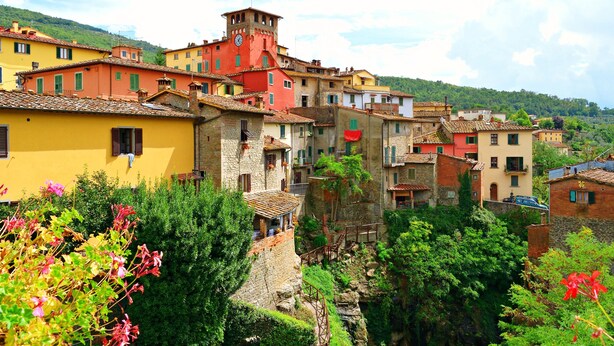 Un poble a la vora d'una collada, amb edificis de colors, una torre destacada i vegetació abundant.