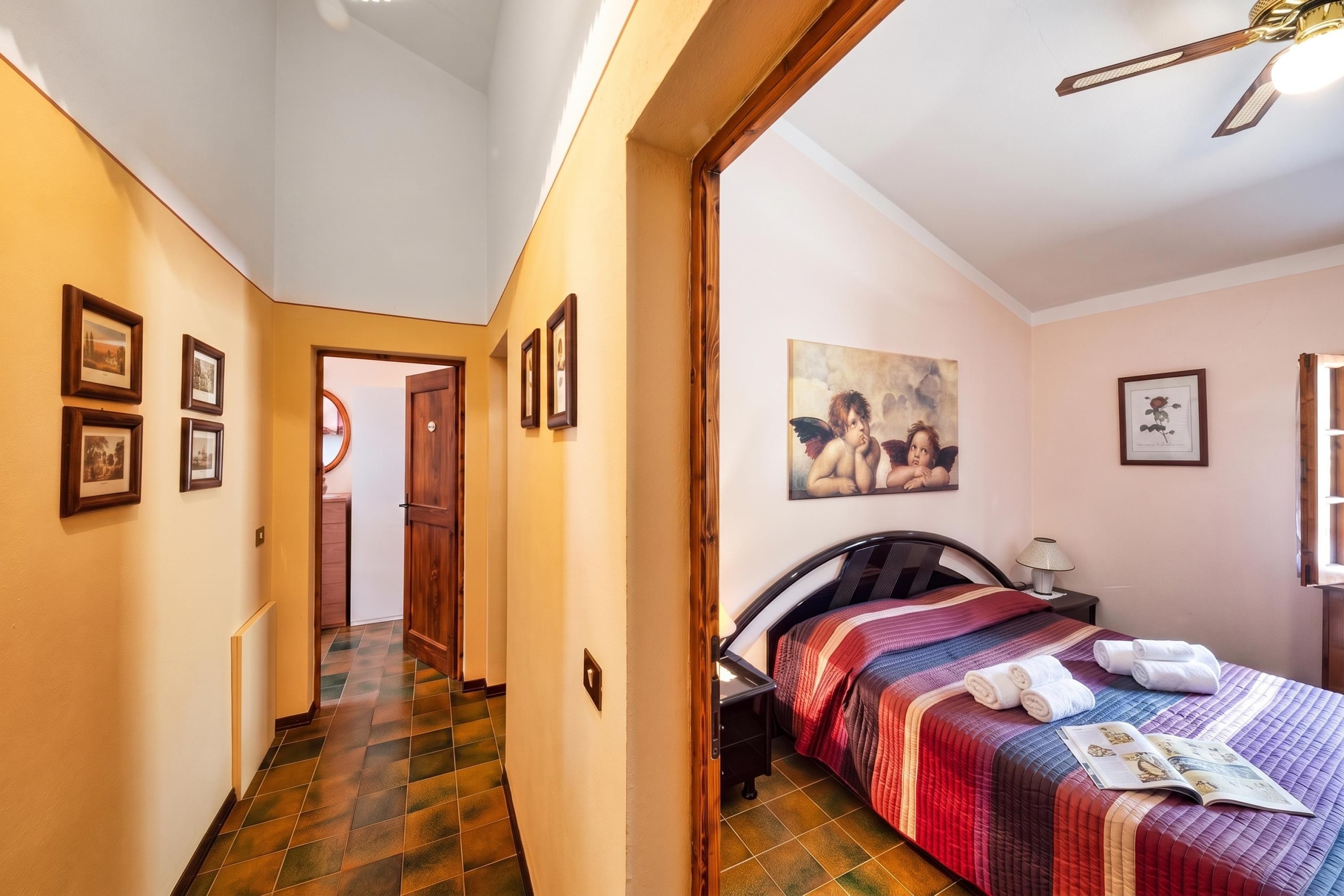 A hotel room with a bed, a bedside table, a nightstand, a lamp, a painting on the wall, and a hallway leading to another room.