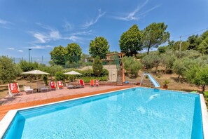 Ein Swimmingpool mit Rutsche, umgeben von Liegestühlen und Sonnenschirmen, mit Bäumen und einem Gebäude im Hintergrund.