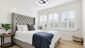 Ein ordentlich eingerichtetes Schlafzimmer mit einer gesteppten Kopfteil, einem Bett mit weißer und dunkelgrauer Bettwäsche, einer Zimmerpflanze und einem Fenster mit weißen Fensterläden.