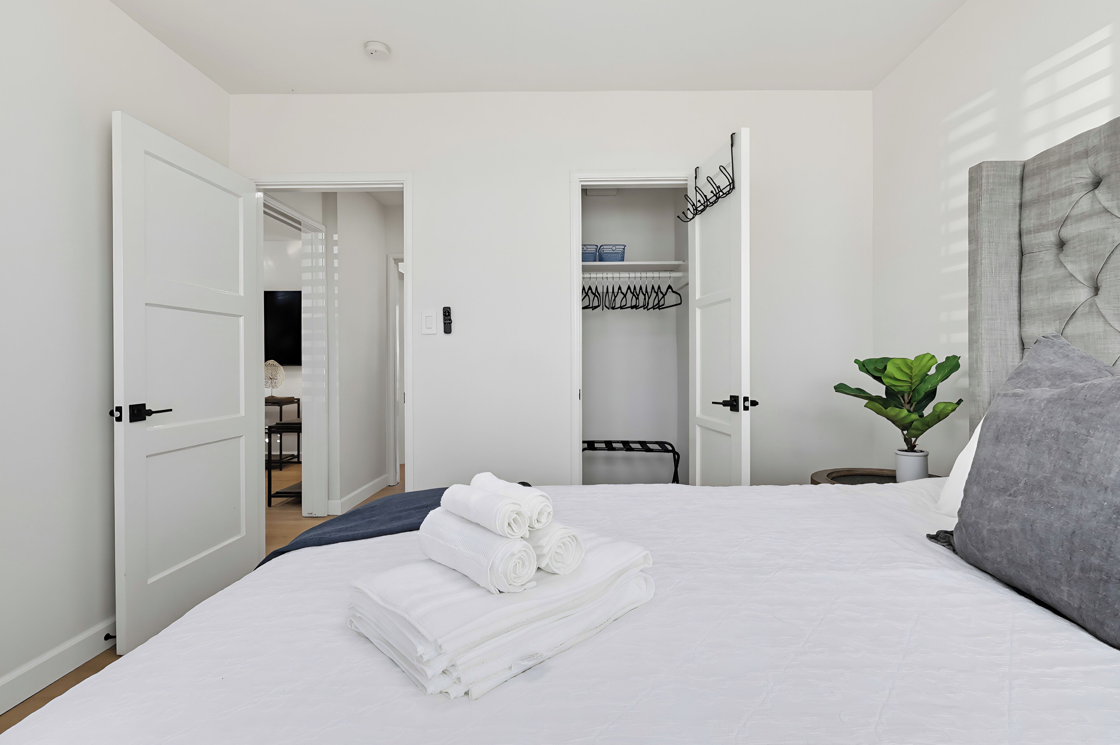 A hotel room with a neatly made bed, a grey upholstered headboard, a potted plant, and a walk-in closet with hanging clothes.