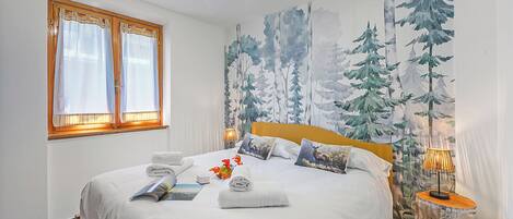 Una cama bien hecha con sábanas blancas, una ventana con cortinas, un mural de árboles en la pared y una lámpara de noche.