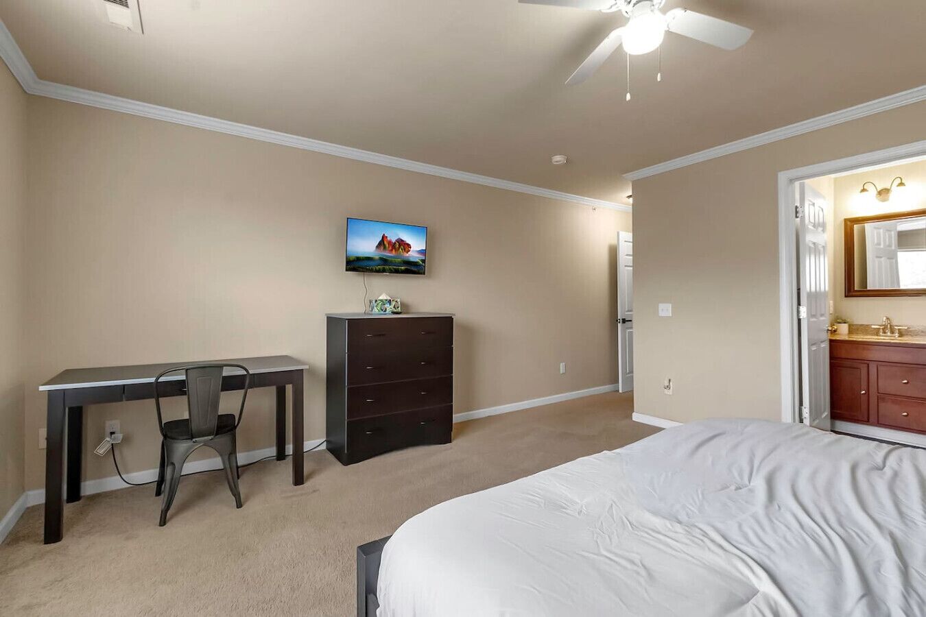 A bedroom with a bed, a desk, a dresser, and a television on the wall.