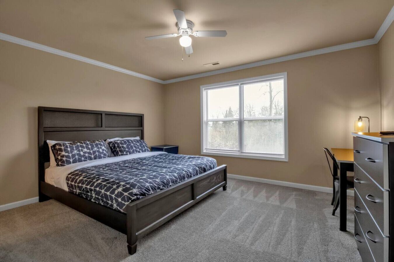 A bedroom with a bed, a desk, a chair, a dresser, a window, and a ceiling fan.