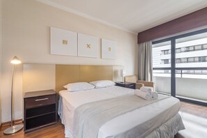Una habitación de hotel con una cama grande, una mesita de noche con lámpara, y vistas a la ciudad a través de un ventanal amplio.