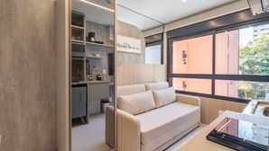 A modern hotel room with a sofa, a desk with a tablet, a coffee machine, and a view of the city through large windows. - Melo Alves 31 2p 1q (São Paulo)