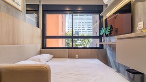 A modern hotel room with a bed, a small seating area, a window with a view of buildings, and a plant on the windowsill. - Melo Alves 31 2p 1q (São Paulo)