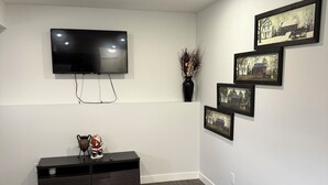 A modern living room with a flat-screen TV, a dark wooden entertainment unit, a glass coffee table, and a wall-mounted fireplace. - RR Luxury Basement Suite, located in a peaceful and friendly neighborhood (Brandon)
