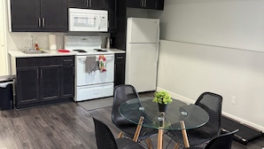A compact kitchen with dark cabinets, a white refrigerator, a microwave, and a stove. There is a glass table with four chairs and a small potted plant. - RR Luxury Basement Suite, located in a peaceful and friendly neighborhood (Brandon)