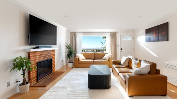 A modern living room with a flat-screen TV, a fireplace, a sofa set, and a large window offering a city view.