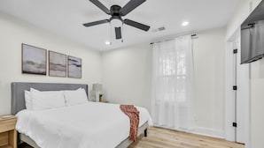Une chambre à coucher avec un lit, un ventilateur de plafond, une table de chevet, une lampe et une fenêtre avec des rideaux.